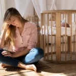 mãe com sinais de depressão pós-parto sentada no chão ao lado do berço do bebê com expressão de cansaço