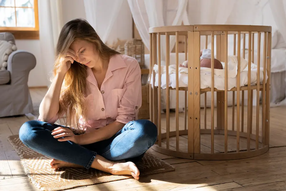 mãe com sinais de depressão pós-parto sentada no chão ao lado do berço do bebê com expressão de cansaço